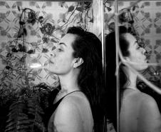foto em preto e branco, mulher encostada em uma parede com um espelho refletindo sua imagem ao lado. ao fundo, plantas na parede decoram o cenário